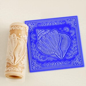 May include: A beige cylindrical rolling pin and a square blue flat plate, both featuring an embossed seashell design surrounded by intricate swirling patterns and decorative borders for creating detailed impressions in soft materials.