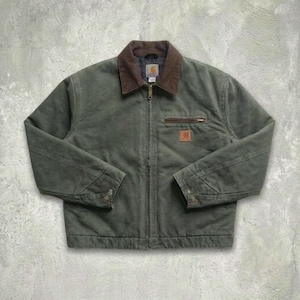 Op de afbeelding: Een donker olijfgroen werkjack met een bruine corduroy kraag, een volledige ritssluiting aan de voorzijde en een borstzak met rits. Een klein vierkant Carhartt-merklabel is zichtbaar op de borstzak en de mouwen hebben verstelbare drukknopen.