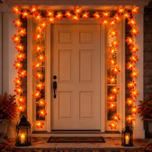 Può includere: Una catena di luci LED con foglie d'acero arancioni e gialle drappeggiata attorno a una porta. Le foglie illuminate creano un bagliore caldo che incornicia l'ingresso. Due lanterne in metallo nero con candele accese sono a terra.