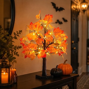 Peut inclure: Un arbre décoratif de table doté de feuilles d'érable artificielles orange et rouges, illuminé par de petites lumières LED blanc chaud. L'arbre possède un tronc texturé brun foncé et une base carrée, posé sur une surface plane.
