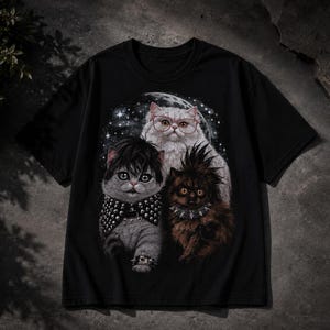 Puede incluir: Una camiseta negra de manga corta con un estampado gráfico de tres gatos. Un gato tiene el pelo oscuro y un collar de tachuelas, otro es blanco con gafas rojas, y el tercero es marrón con una cresta y un collar de pinchos sobre un cielo estrellado.