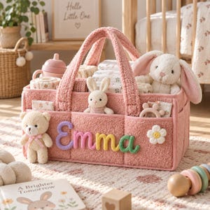 May include: A pink textured fabric storage basket featuring the name Emma in colorful yarn letters. The basket contains a plush teddy bear, a small crocheted rabbit, a larger plush rabbit, a baby bottle, and a white flower applique on the front.