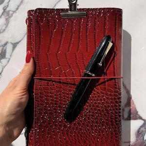 May include: A deep red notebook with a glossy crocodile-embossed texture, secured by a thin red elastic band. A silver metal clip is attached to the top edge, and a black fountain pen with gold-tone accents rests diagonally across the front cover.