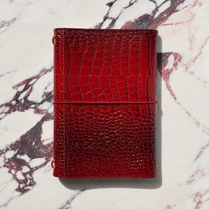 May include: A rectangular notebook cover in a deep red color with a glossy, textured crocodile skin pattern. The cover features a thin, matching red elastic band wrapped horizontally around the center to keep the notebook securely closed.