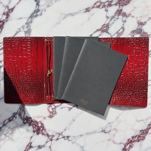 May include: A red faux-leather journal cover with a textured crocodile skin pattern, featuring gold-toned elastic cords for holding inserts. Inside are three black notebooks with stitched spines, and the front notebook has the word "naked" in gold.