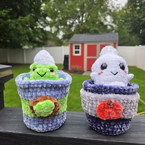 May include: Two plush crochet figures in textured yarn pots. The left pot is blue and green with a green frog wearing a white hat. The right pot is dark blue and gray with a white ghost wearing a white hat and a small orange pumpkin accent.