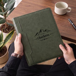 May include: A dark olive green leather portfolio folder featuring a black engraved mountain graphic and the name Charlotte centered on the front cover. The rectangular folder has a smooth, matte finish and a vertical stitched seam detail on the left side.