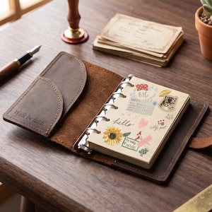 May include: A brown leather ring-bound planner with a pocket on the left cover embossed with the text Walk by faith. The interior pages feature various stickers including a sunflower, a cat, a habit tracker, and the handwritten word hello.