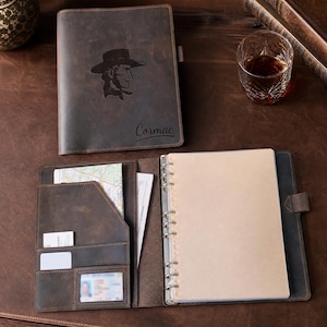 May include: A dark brown leather binder with a cowboy silhouette engraving and the name Cormac. The interior features multiple pockets for cards and documents, a clear ID window, and a six-ring metal mechanism holding blank paper pages.