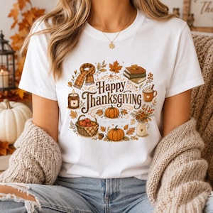May include: A white t-shirt featuring a colorful graphic print with the text Happy Thanksgiving. The design includes illustrations of a plaid scarf, stacked books, a candle, a mug, a basket of apples, pumpkins, and autumn leaves.