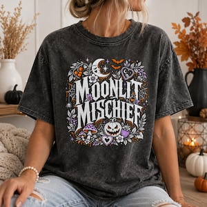 May include: A charcoal grey mineral wash t-shirt featuring a graphic print with the text MOONLIT MISCHIEF. The design includes a crescent moon, bats, a jack-o'-lantern, mushrooms, spiderwebs, and floral elements in shades of orange, purple, and white.