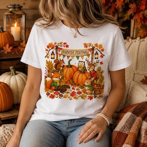 May include: A white t-shirt featuring a colorful graphic of woodland animals including a squirrel, fox, hedgehog, owl, and deer surrounding pumpkins and autumn leaves with a banner that reads Harvest Festival in brown lettering.