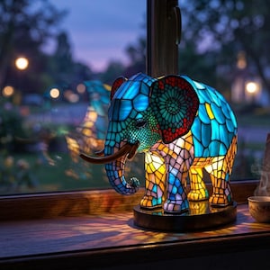 Op de afbeelding: Een glas-in-loodlamp in de vorm van een olifant met een mozaïekpatroon van blauwe, gele en rode glasstukjes. De lamp heeft gebogen bruine slagtanden, staat op een donkere ronde voet en verspreidt een warme gloed vanuit het kleurrijke lichaam.
