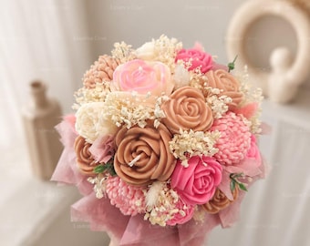 May include: A bouquet of wax candles shaped like roses and other flowers in shades of pink, beige, and cream, accented with dried white baby's breath and wrapped in translucent pink paper, featuring visible candle wicks in the center of the flowers.