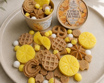 May include: A collection of wax melts shaped like citrus slices, waffles, pretzels, and cookies, with a paper container labeled Essence Cove Citrus Cookie Crumble. The wax pieces are yellow and tan, accompanied by small white and yellow wax dollops.