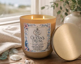 May include: A two-wick candle in a frosted tan glass jar with a gold lid. The label reads The Ocean's Muse, Ocean Breeze, Hibiscus, Soft Musk, Sandalwood, Luxury Olive Oil Wax Candle, Net Wt. 10 oz (283 g). The candle is lit and glowing.
