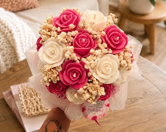 May include: A bouquet of rose-shaped candles in shades of deep pink and cream, interspersed with dried baby's breath flowers. The arrangement is wrapped in translucent white paper and features a circular tag with the text Pink Spell.