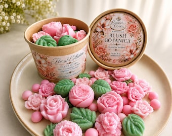 May include: A cardboard container and lid labeled Essence Cove Blush Botanica Coconut Soy Wax Melts, filled with pink rose-shaped and green leaf-shaped wax pieces. Additional pink and green wax shapes are scattered on a plate in front of the container.