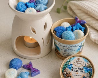 May include: A cream-colored ceramic wax warmer with a lit tea light candle inside, accompanied by a cardboard container and lid labeled Coastal Collection Sea Side Treasures, filled with blue, white, and purple seashell-shaped wax melts.