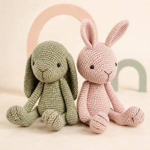 Puede incluir: Dos animales de peluche tejidos a mano en forma de conejos. Un conejo es de color verde salvia con orejas largas y caídas, y el otro es de color rosa claro con orejas erguidas. Ambos tienen ojos de cuentas negras y narices bordadas.