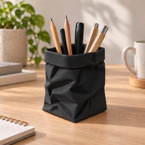 Könnte beinhalten: Ein schwarzer, flexibler, stoffartiger Schreibtisch-Organizer mit umgeschlagenem oberen Rand, der eine Sammlung von Schreibgeräten enthält, darunter Holzstifte, schwarze Stifte und Stifte mit Metallkappen, die aufrecht stehen.