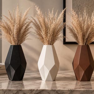 Puede incluir: Tres floreros geométricos en negro, blanco y marrón, cada uno con una superficie facetada y angular. Cada florero contiene un arreglo alto de hierba de la pampa beige, seca y plumosa, creando una exhibición uniforme de tres recipientes.