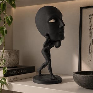 Pode incluir: Uma escultura em preto fosco representando uma figura musculosa caminhando para frente enquanto carrega uma cabeça humana grande e sobredimensionada sobre os ombros. A figura está posicionada sobre uma base circular de acabamento escuro.