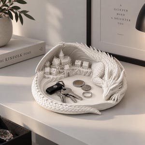 May include: A white circular trinket tray featuring a sculpted dragon curled around a miniature village with houses and a castle. The dragon's tail and wings form the outer edge of the tray, which holds a set of metal keys and several coins.