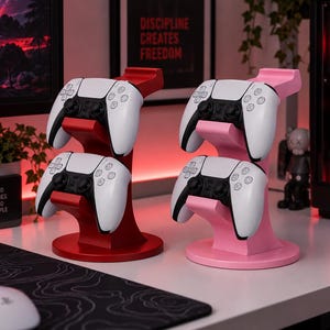 Op de afbeelding: Twee verticale controllerstandaards, één rood en één roze, die elk twee wit-zwarte gamecontrollers vasthouden. De standaards hebben een ronde voet en twee trapsgewijze houders. De controllers zijn voorzien van analoge sticks en knoppen.