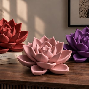Op de afbeelding: Drie keramische vetplantsculpturen in rood, lichtroze en paars. Het rode beeld staat op twee gestapelde boeken met de titels The KINFOLK Home en A Life More Considered. Elk stuk heeft gelaagde, puntige bloemblaadjes in een bloeiende vorm.