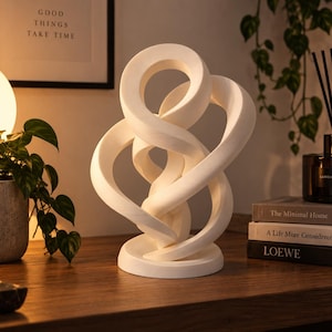 May include: A white abstract sculpture featuring a series of interconnected, smooth, curved loops that form a continuous, flowing structure. The piece is mounted on a circular base and has a matte finish, creating a balanced and complex geometric form.