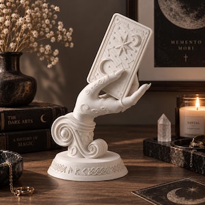 Puede incluir: Un soporte escultórico de mano blanca con volutas intrincadas y una base circular con símbolos rúnicos grabados. La mano está posicionada para sostener una tarjeta rectangular con motivos celestiales en relieve, incluyendo una estrella grande, una luna creciente y constelaciones.