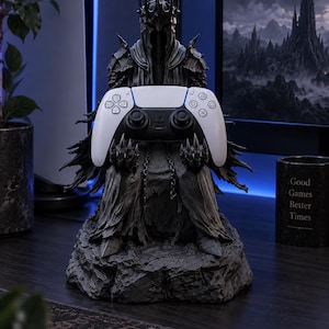 May include: A dark gray, textured resin statue of a hooded, armored figure with a spiked crown, seated on a rocky base. The figure holds a white and black wireless game controller in its hands. The statue features intricate, jagged armor details.