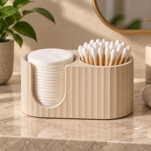 May include: A beige, oval-shaped bathroom organiser with a vertical ribbed texture. The container features two distinct compartments: one open-front section holding a stack of white cotton rounds and a second cylindrical section filled with cotton swabs.