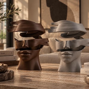 Op de afbeelding: Twee abstracte hoofdsculpturen, één in donkerbruin en één in lichtgrijs, met een gefragmenteerd ontwerp waarbij het bovenste deel van het gezicht en het voorhoofd door een horizontale opening in het materiaal van de neus en lippen zijn gescheiden.