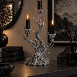 May include: A grey resin candelabra featuring a central human skull base surrounded by sculpted tree branches and leaves. The structure holds three lit black pillar candles in gothic-style holders, creating a dark, textured decorative centrepiece.