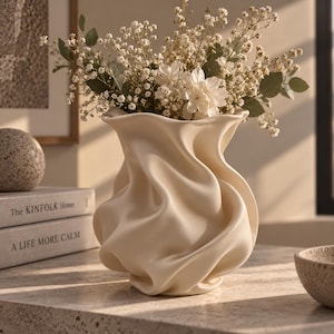 Pode incluir: Um vaso de cerâmica na cor creme com uma textura distintamente ondulada, fluida e drapeada. O vaso apresenta uma borda larga e sinuosa e um corpo arredondado, preenchido com um buquê de pequenas flores brancas e caules de folhagem verde.