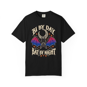 May include: A black t-shirt featuring a graphic of a bat with wings colored in pink, purple, and blue. Above the bat is a crescent moon, and below it are small orange leaves. The text reads "BI BY DAY" at the top and "BAT BY NIGHT" at the bottom.