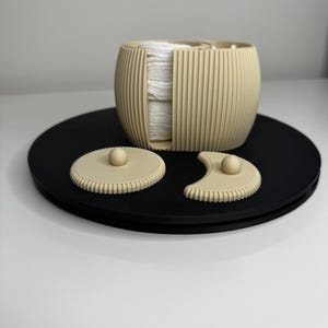 Può includere: Un contenitore cilindrico beige a coste con una fessura verticale che contiene una pila di dischetti di cotone bianchi. Due coperchi separati, uno circolare e uno a forma di mezzaluna, dotati di un piccolo pomello, sono posti davanti.