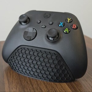 May include: A black video game controller featuring a matte finish, two analogue sticks, a directional pad, and four action buttons labelled Y, X, A, and B in yellow, blue, green, and red. The lower front section has a prominent black honeycomb texture.