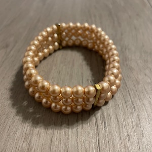 Può includere: Un bracciale a tre fili composto da perle artificiali rotonde color pesca. I tre fili sono tenuti insieme da due distanziatori metallici verticali e rettangolari color oro, posizionati sui lati opposti della struttura circolare.