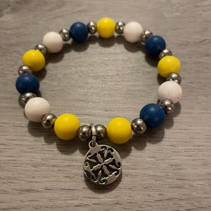 May include: A stretch bracelet featuring alternating matte yellow, navy blue, and white spherical beads interspersed with smaller metallic silver-toned beads. A circular silver-toned metal charm with an intricate cut-out floral pattern hangs from the center.