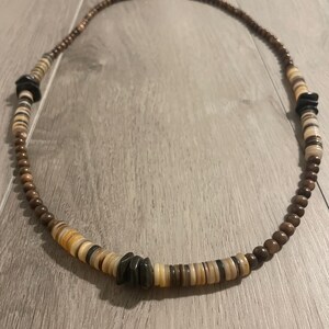 May include: A long necklace featuring a repeating pattern of small brown spherical beads, flat circular disc beads in shades of cream, tan, and brown, and irregular dark charcoal gray stone-like accents arranged symmetrically along the strand.
