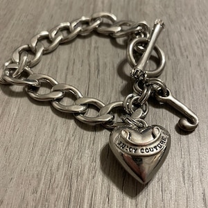 May include: A silver-toned chain link bracelet featuring a toggle clasp closure. The bracelet includes a heart-shaped charm engraved with the text Juicy Couture and a separate J-shaped charm hanging from the circular toggle ring component.