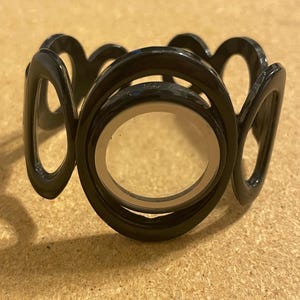 May include: A black cuff bracelet featuring a series of interconnected circular and oval shapes. The central element is a large black circle with a clear, transparent center, surrounded by a repeating pattern of open, rounded geometric links.
