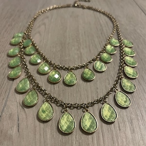 May include: A two-layered gold-tone chain necklace featuring multiple dangling, teardrop-shaped green faceted stones. The necklace has a metallic finish and a clasp closure at the back, with the stones arranged in a symmetrical pattern along the chains.