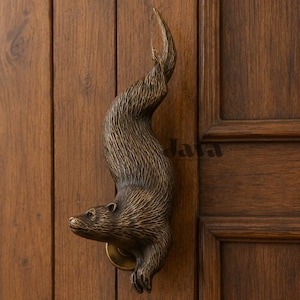 May include: A bronze-toned metal door handle shaped like a swimming otter with detailed fur texture. The otter is oriented vertically with its head pointing downward and its tail curving upward. The handle is mounted onto a dark brown wooden door.
