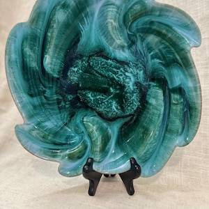 May include: A decorative glass bowl featuring a swirling pattern of deep forest green, teal, and white colors. The bowl has a scalloped, wavy edge and a textured, dark green center. It is displayed on a small, black plastic tripod stand.
