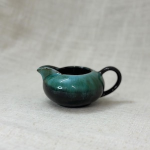 Peut inclure: Un petit pot à lait en céramique avec un vernis brillant passant d'une base noire profonde à une section supérieure turquoise et bleu sarcelle. Le récipient présente un bec verseur incurvé et une anse noire unique sur le côté.