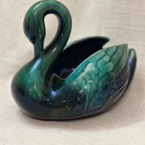 Peut inclure: Un objet en céramique en forme de cygne, servant de cache-pot ou de coupelle, doté d'une glaçure brillante aux nuances variées de vert émeraude, vert forêt et bleu foncé. Le cygne présente un cou courbé et des détails de plumes moulés.